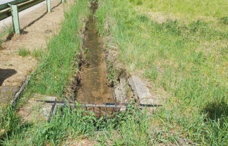 Hahnwiesenbach – Gerader Bachverlauf mit Sohlverbau Bachlauf zwischen Wiesen mit Gras und einem Metallgitter